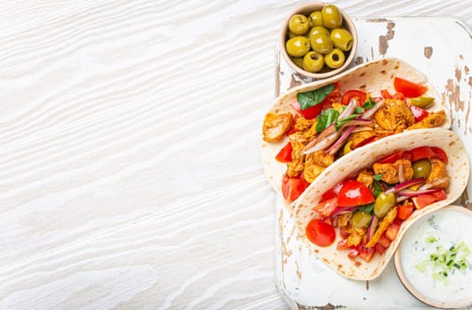 Traditional Greek Dish Gyros: Pita bread Wraps with vegetables, meat, herbs, olives on rustic wooden cutting board with Tzatziki sauce, olive oil top view on white wooden background, space for text.