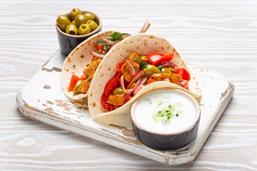 Traditional Greek Dish Gyros: Pita bread Wraps with vegetables, meat, herbs, olives on rustic wooden cutting board with Tzatziki sauce, olive oil top view, white wooden background, angle view.