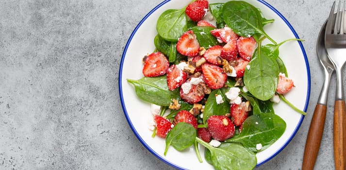 Light Healthy Summer Salad with fresh Strawberries, Spinach, Cream Cheese and Walnuts on White Ceramic Plate, Grey rustic stone Background Top View Copy Space.