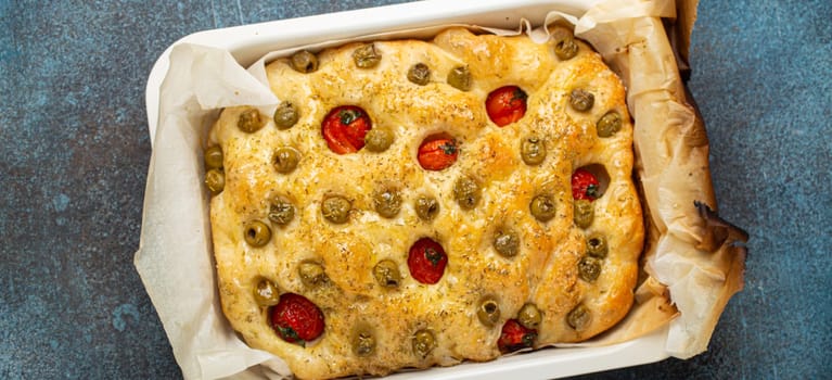 Overhead of Traditional Italian Homemade Flat Bread Focaccia with Green Olives, Olive Oil, Cherry Tomatoes and Rosemary in Baking Tray on Rustic Dark Blue Concrete Background