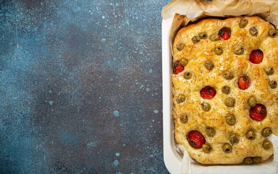 Overhead of Traditional Italian Homemade Flat Bread Focaccia with Green Olives, Olive Oil, Cherry Tomatoes and Rosemary in Baking Tray on Rustic Dark Blue Stone Background, Space for Text