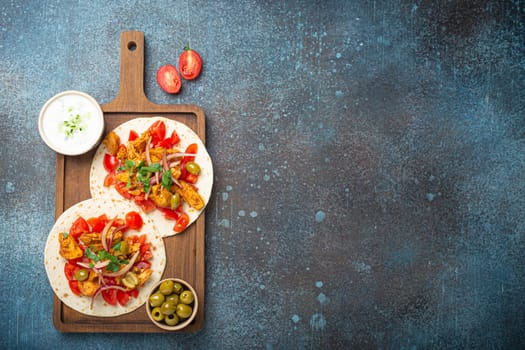 Cooking Traditional Greek Dish Gyros: Pita bread with vegetables, meat, herbs, olives on rustic wooden cutting board with Tzatziki sauce, olive oil top view, dark blue stone background, copy space.