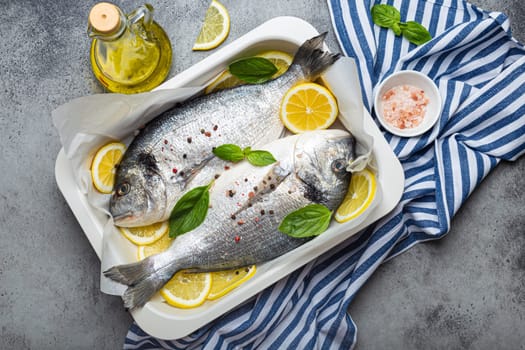 Raw fish dorado in casserole dish with ingredients lemon, fresh basil, bottle of olive oil on wooden cutting board with knife on rustic stone background top view, cooking healthy fish dorado concept