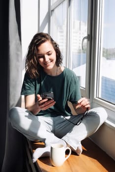 Lockdown Lifestyle: Young Woman Using Smartphone at Home, Engaging in Relaxation and Connection While Looking Out of Window - Technology in Leisure Time.