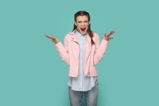 Portrait of angry aggressive teenager girl with braids wearing pink jacket standing with raised arms, screaming with hate. Indoor studio shot isolated on green background.