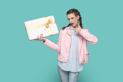 Portrait of joyful flirting teenager girl with braids wearing pink jacket standing and pointing at gift box, winking to camera. Indoor studio shot isolated on green background.