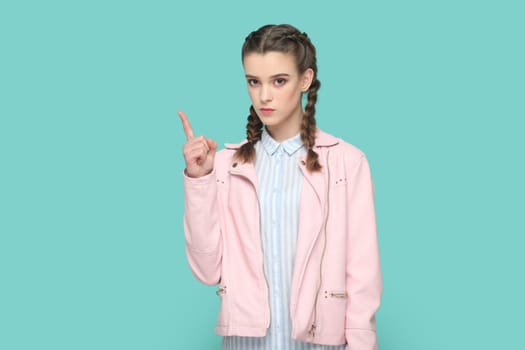 Portrait of serious bossy teenager girl with braids wearing pink jacket standing with raised finger, warning her classmates about danger. Indoor studio shot isolated on green background.