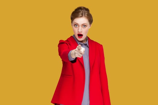 Portrait of shocked astonished woman with red lips standing indicating fore finger to you, being amazed and surprised, choosing you, wearing jacket. Indoor studio shot isolated on yellow background.