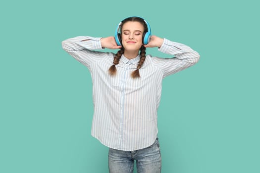 Portrait of teenager girl with braids wearing striped shirt standing listening favorite music with headphones, closed eyes and smiling. Indoor studio shot isolated on green background.