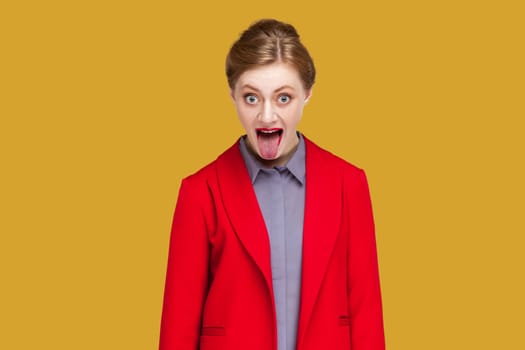 Portrait of woman with red lips sticks out tongue, makes happy grimace, has long hair, expresses positive emotions, wearing red jacket. Indoor studio shot isolated on yellow background.