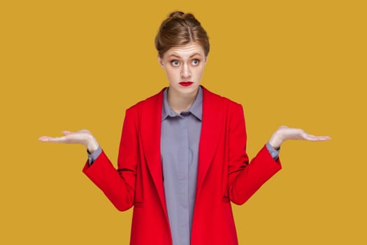 I do not know. Puzzled uncertain woman with red lips standing and spreads hands, looking at camera with confused expression, wearing red jacket. Indoor studio shot isolated on yellow background.