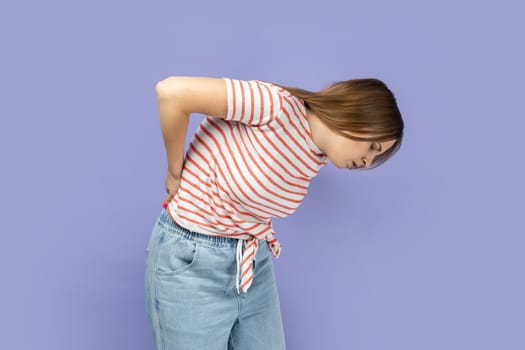 Portrait of unhealthy sick ill blond young adult woman wearing striped T-shirt having discomfort lower lumbar muscular, kidney stones. Indoor studio shot isolated on purple background.