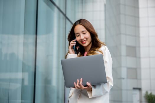 Portrait beautiful business woman smiling holding computer discussing issues on smart mobile phone in city, Asian businesswoman working on laptop and talking cell phone at front building near office