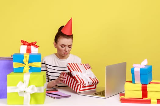 Unhappy woman in party cone sitting in office workplace, opening gift box and looking inside with sad upset expression, unwrapping present. Indoor studio studio shot isolated on yellow background