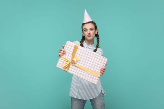 Portrait of funny positive teenager girl with braids wearing striped shirt and party cone, holding gift in wrapped box, looks at camera with pout lips. Indoor studio shot isolated on green background.