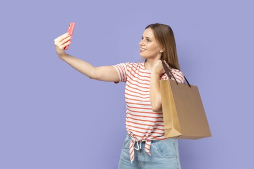 Portrait of young adult blond woman blogger wearing striped T-shirt standing holding shopping bag and making selfie or broadcasting livestream. Indoor studio shot isolated on purple background.