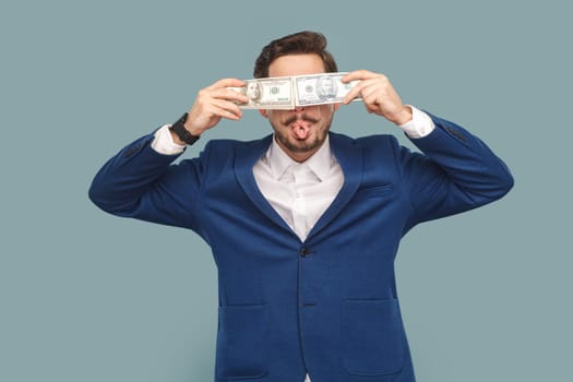 Funny positive childish man with mustache standing showing tongue out and covering eyes with dollar banknotes, wearing official style suit. Indoor studio shot isolated on light blue background.