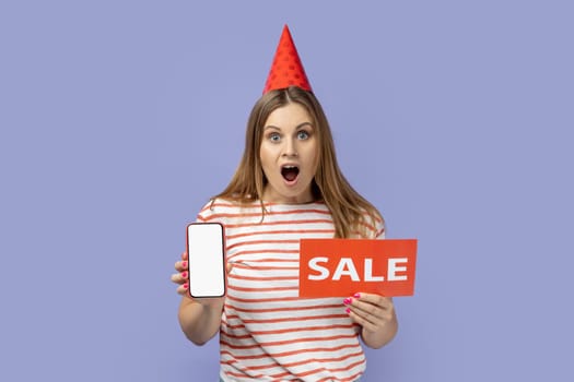 Portrait of shocked amazed blond woman wearing striped T-shirt and party cone holding mobile phone with empty screen and car with sale inscription. Indoor studio shot isolated on purple background.