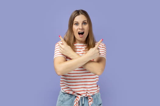 Portrait of amazed shocked woman wearing striped T-shirt standing with crossed hands and pointing to empty space for promotional text on both sides. Indoor studio shot isolated on purple background.