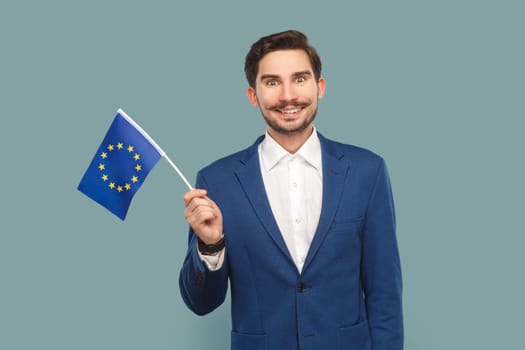 Portrait of smiling happy handsome man with mustache standing waving europe union flag, expressing happiness, wearing white shirt and jacket. Indoor studio shot isolated on light blue background.