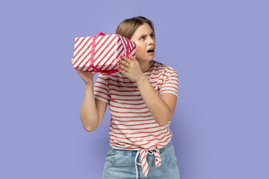 Portrait of thoughtful curious blond woman wearing striped T-shirt shaking gift box and looking away, trying to guess what inside in present box. Indoor studio shot isolated on purple background.