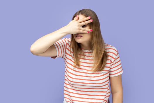 Young curious blond woman wearing striped T-shirt looking through fingers, peeking with serious calm face, having suspicious, watching secrets. Indoor studio shot isolated on purple background.