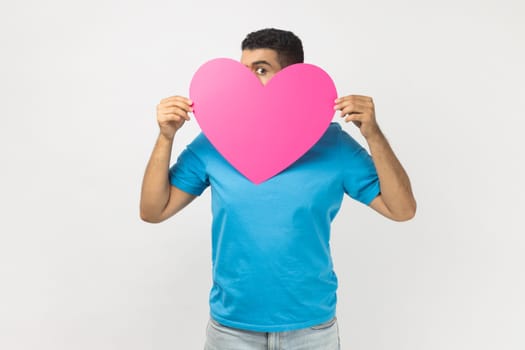 Portrait of shy anonymous man wearing blue T- shirt standing peeking from big pink heart, being shy to express his feelings on Valentines day. Indoor studio shot isolated on gray background.