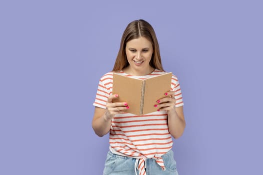 Portrait of satisfied smiling woman wearing striped T-shirt holding and reading book, being very interested in plot, expressing positive emotions. Indoor studio shot isolated on purple background.