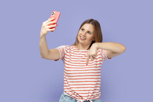 Portrait of adorable positive blond woman in T-shirt standing with mobile phone and broadcasting livestream, pointing down, asking to subscribe. Indoor studio shot isolated on purple background.