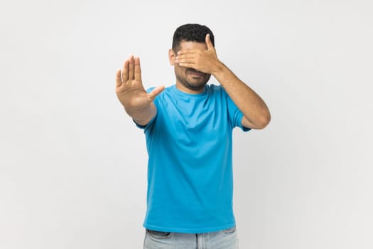 Portrait of young adult unshaven man wearing blue T- shirt standing covering his face with hand, hiding his face, making stop gesture. Indoor studio shot isolated on gray background.