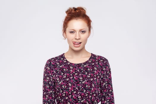Portrait of flirting positive optimistic redhead woman with bun hairstyle wearing dress standing and showing tongue out, winking to camera. Indoor studio shot isolated on gray background.