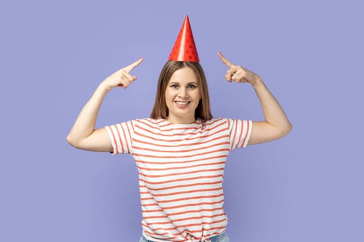 Portrait of blond woman wearing striped T-shirt pointing to funny party cone on head and smiling to camera, celebrating career growth. Indoor studio shot isolated on purple background.