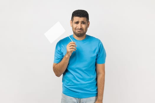 I give up. Portrait of unshaven man wearing blue T- shirt standing and holding white flag and looking at camera with sad face, crying. Indoor studio shot isolated on gray background.