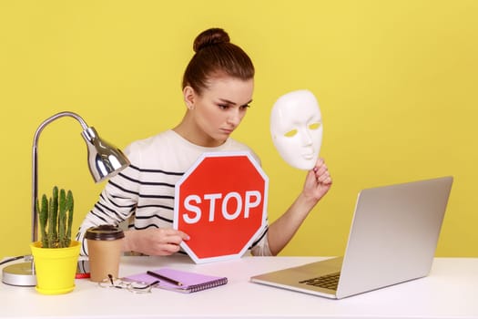 Portrait of serious young adult woman holding white mask with unknown face and red traffic sign while at laptop display. Indoor studio studio shot isolated on yellow background.