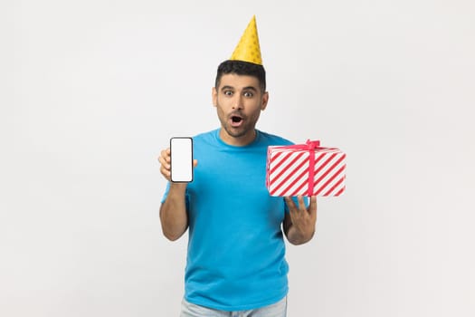 Shocked astonished surprised unshaven man wearing blue T- shirt and yellow party cone holding present box and showing cell phone with empty display. Indoor studio shot isolated on gray background.