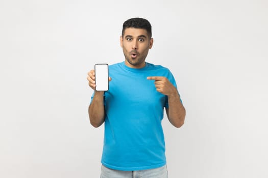 Portrait of shocked amazed unshaven man wearing blue T- shirt standing pointing at smart phone with blank white display, mockup for promotional text. Indoor studio shot isolated on gray background.