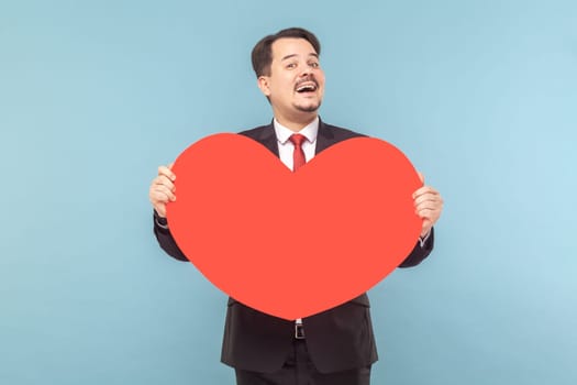 Portrait of satisfied joyful man with mustache standing holding big red card, expressing love and gentle, wearing black suit with red tie. Indoor studio shot isolated on light blue background.