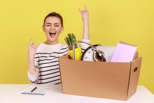 Optimistic woman office worker sitting at workplace with cardboard box with her things and yelling, showing victory sign, rejoice a new job. Indoor studio studio shot isolated on yellow background.