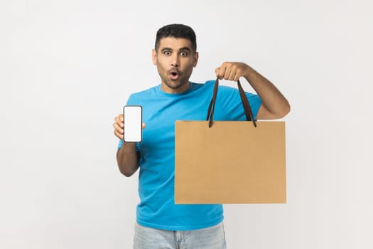 Portrait of shocked astonished handsome man wearing blue T- shirt standing holding shopping bag and mobile phone with blank screen for advertisement. Indoor studio shot isolated on gray background.