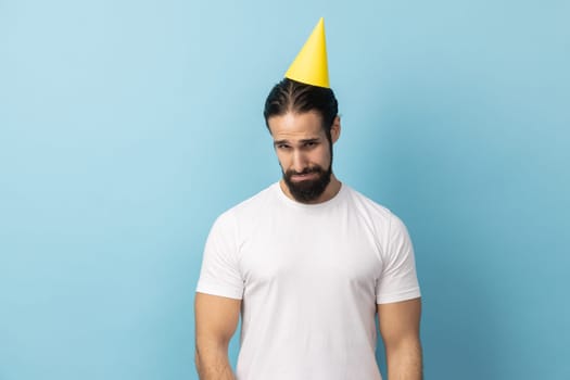 Portrait of sad unhappy man with beard wearing white T-shirt and party cone standing alone, nobody came to his birthday party, expressing sadness. Indoor studio shot isolated on blue background.