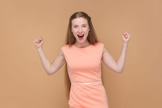 Portrait of extremely happy positive woman with long hair standing with clenched fists, screaming with excitement, wearing elegant dress. Indoor studio shot isolated on brown background.