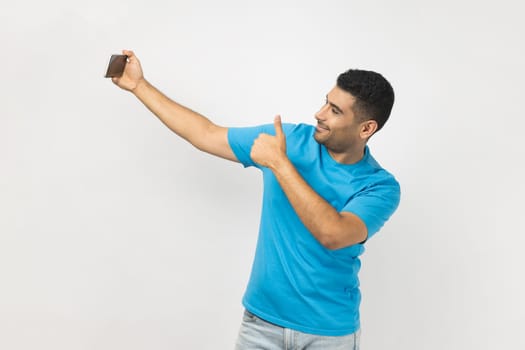 Portrait of joyful satisfied delighted man blogger in blue T- shirt holding mobile phone, has video call or livestream, showing thumb up, like gesture. Indoor studio shot isolated on gray background.