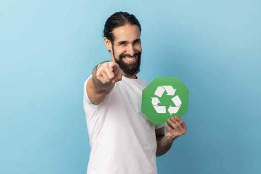 Portrait of man with beard wearing white T-shirt holding green recycling sign, pointing to camera, calls on to save our planet from pollution. Indoor studio shot isolated on blue background.