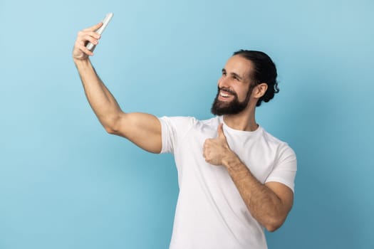 Portrait of man with beard wearing white T-shirt showing thumbs up like gesture and winking looking at phone camera, making selfie or recording video. Indoor studio shot isolated on blue background.