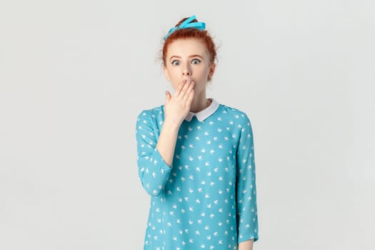 Portrait of shocked astonished adorable ginger woman with bun hairstyle, closed her mouth with palm, hearing secret, wearing blue dress. Indoor studio shot isolated on gray background.