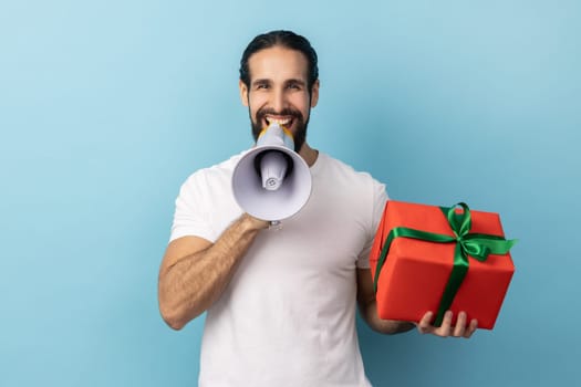 Portrait of positive handsome man with beard wearing white T-shirt holding red present box and screaming in megaphone, making announcement. Indoor studio shot isolated on blue background.