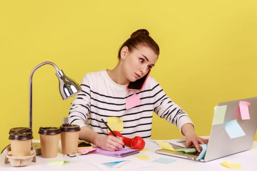Concentrated secretary in striped shirt receiving calls on phone and making notes, writing down information, sitting at workplace with laptop. Indoor studio studio shot isolated on yellow background.