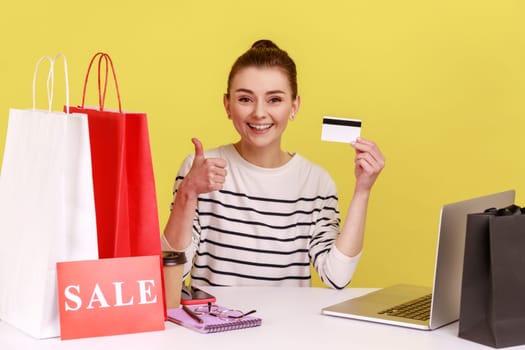 Smiling happy satisfied woman manager sitting at workplace with shopping bags and sale card, showing credit card and thumb up. Indoor studio studio shot isolated on yellow background.