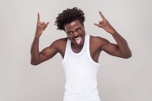 Man with Afro hairstyle shouting from happiness, has widely opened mouth, makes rock n roll gesture, says rock this party. wearing white T-shirt. Indoor studio shot isolated on gray background.