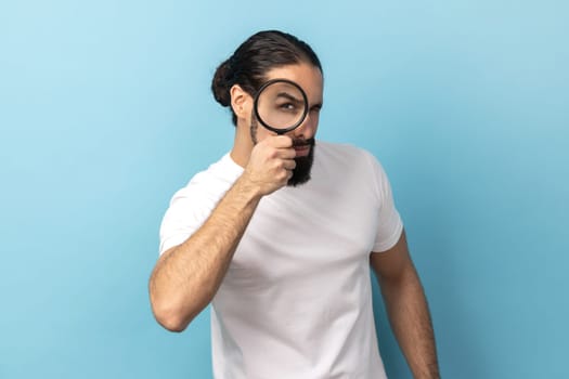 Portrait of man with beard wearing white T-shirt looking through magnifying glass, spying, finding out something, exploring crime scene, inspecting. Indoor studio shot isolated on blue background.